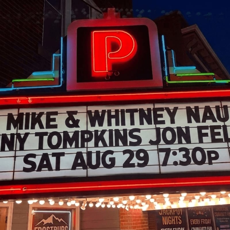 A neon "P" sign illuminates a marquee advertising a comedy show with performers and a date, set against a dark, building-lined street.