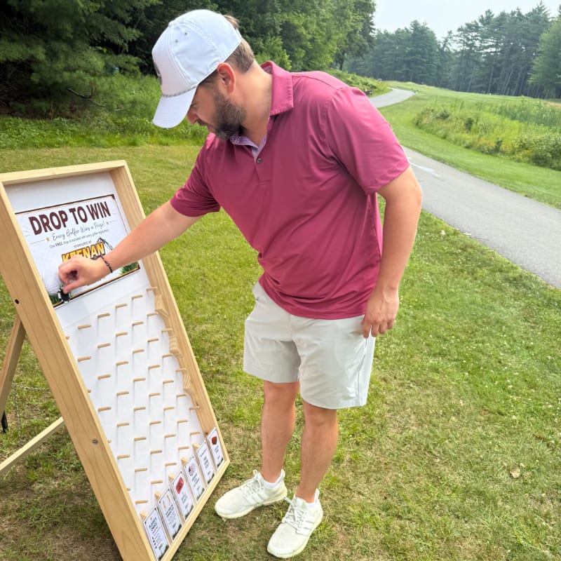 A man in a pink shirt and white hat drops a token into a game board in the foreground, with a golf course and trees in the background.