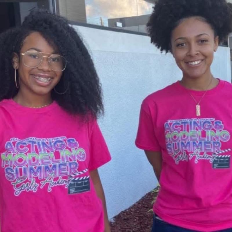 Two young women in matching pink t-shirts stand in front of a textured white wall.