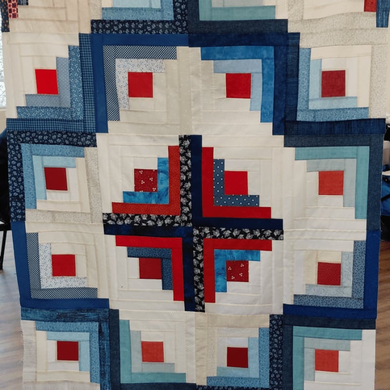 A close-up view of a red, white, and blue log cabin quilt with a central starburst pattern is displayed against a blurred background of furniture and a window.