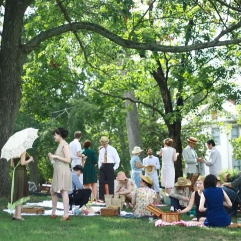 A group of people in vintage attire enjoy a picnic on a grassy lawn in front of a large white house, with lush green trees forming the background.