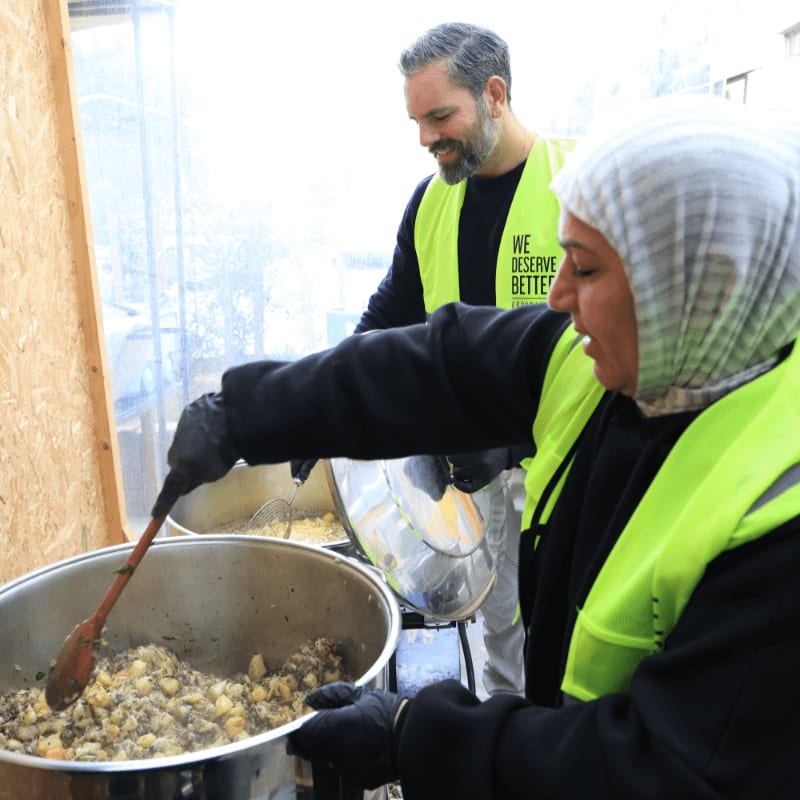 A woman in a hijab stirs food in a large pot in the foreground, while a man in a yellow vest stands behind her in the background.