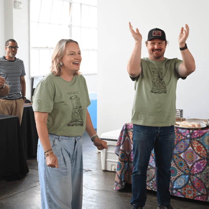 A woman and a man in matching t-shirts clap their hands in front of a table with snacks, while another man stands in the background.