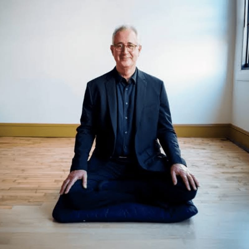 A man in a suit sits cross-legged on a cushion in a room with wooden floors and white walls.