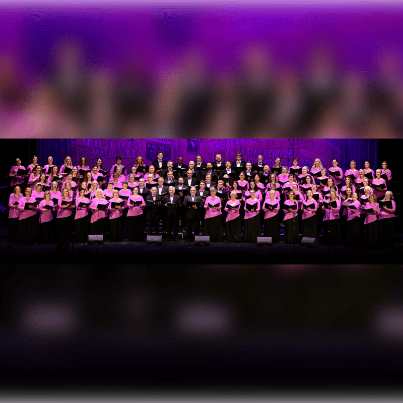 A large choir in pink tops and black bottoms stands on a stage with a purple, blurred background.