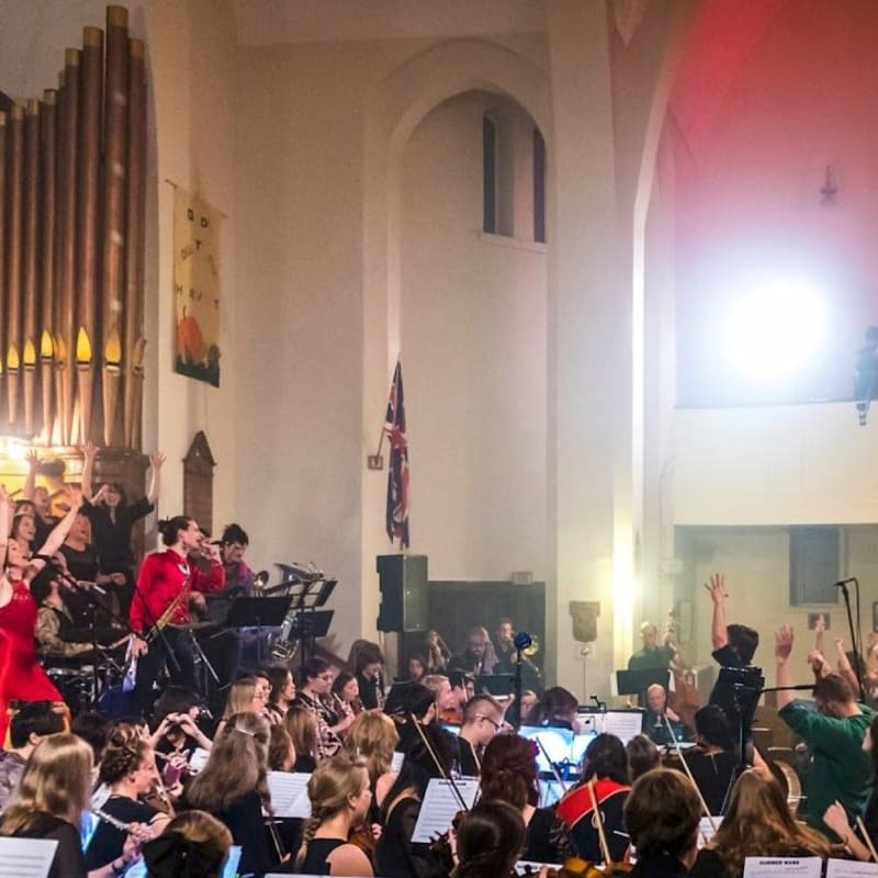 A choir and orchestra perform in a church with tall arched windows and a pipe organ in the background.