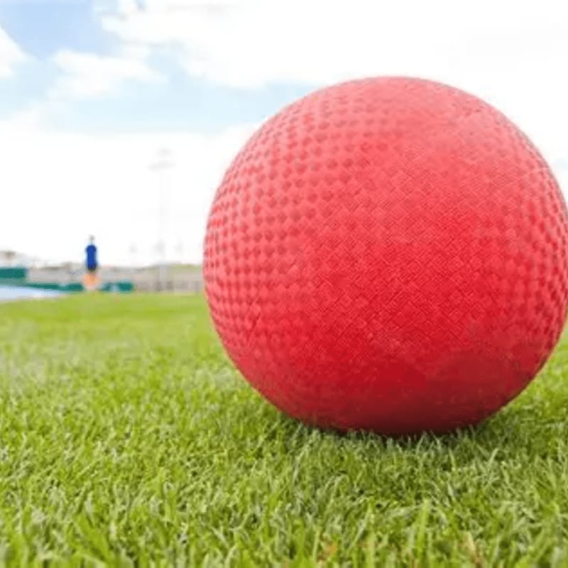 A textured red ball rests on a vibrant green lawn, with a blurred figure and a bright sky in the distance.