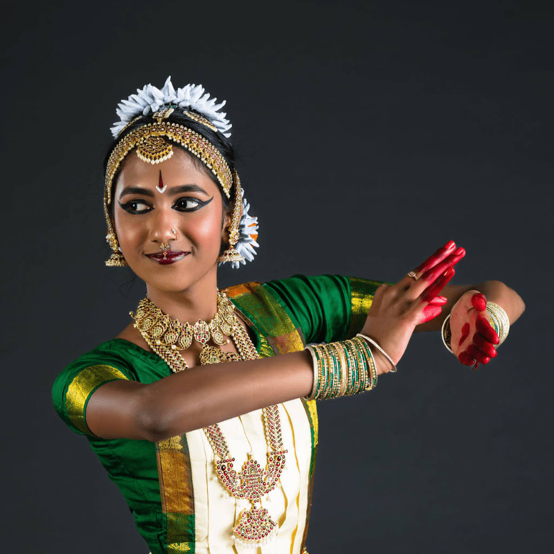 A young woman in traditional Indian dance attire with intricate jewelry and makeup performs a graceful pose against a plain dark background.