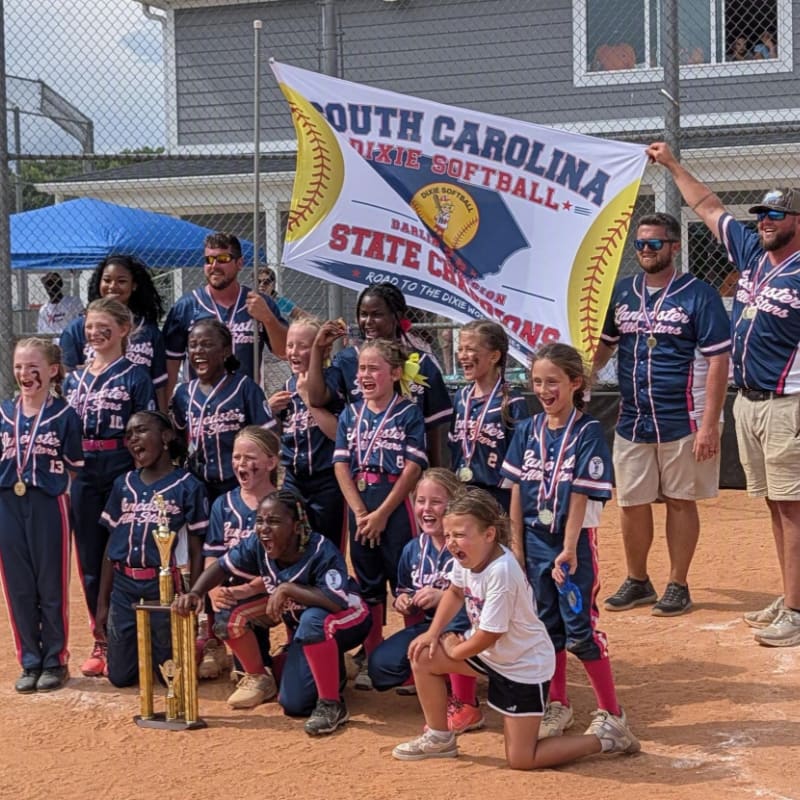 A group of young girls in softball uniforms, some holding a trophy, pose for a photo in front of a banner celebrating their state championship win, with spectators and a building visible in the background.