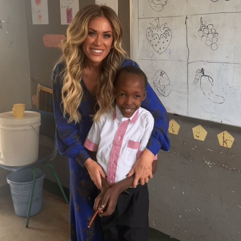 A smiling woman in a blue dress embraces a young boy in a school uniform in front of a whiteboard with children's drawings.