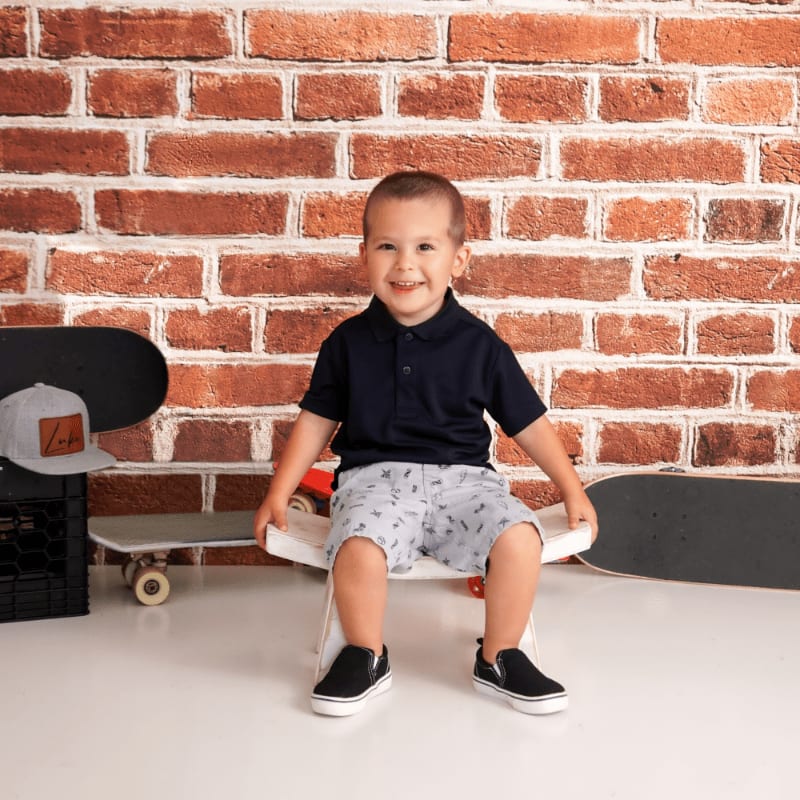A smiling young boy in a navy polo and patterned shorts sits on a skateboard in front of a brick wall, with other skateboards and a crate to his sides.