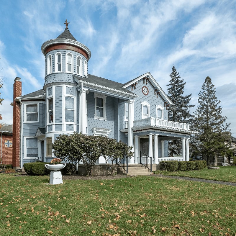 A light blue Victorian house with a prominent turret stands on a green lawn scattered with fallen leaves, under a bright blue sky with wispy clouds.