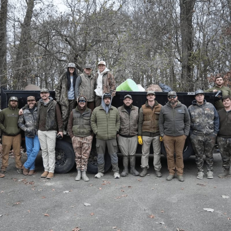 A group of people in hunting attire stand in front of a truck, with a wooded area in the background.