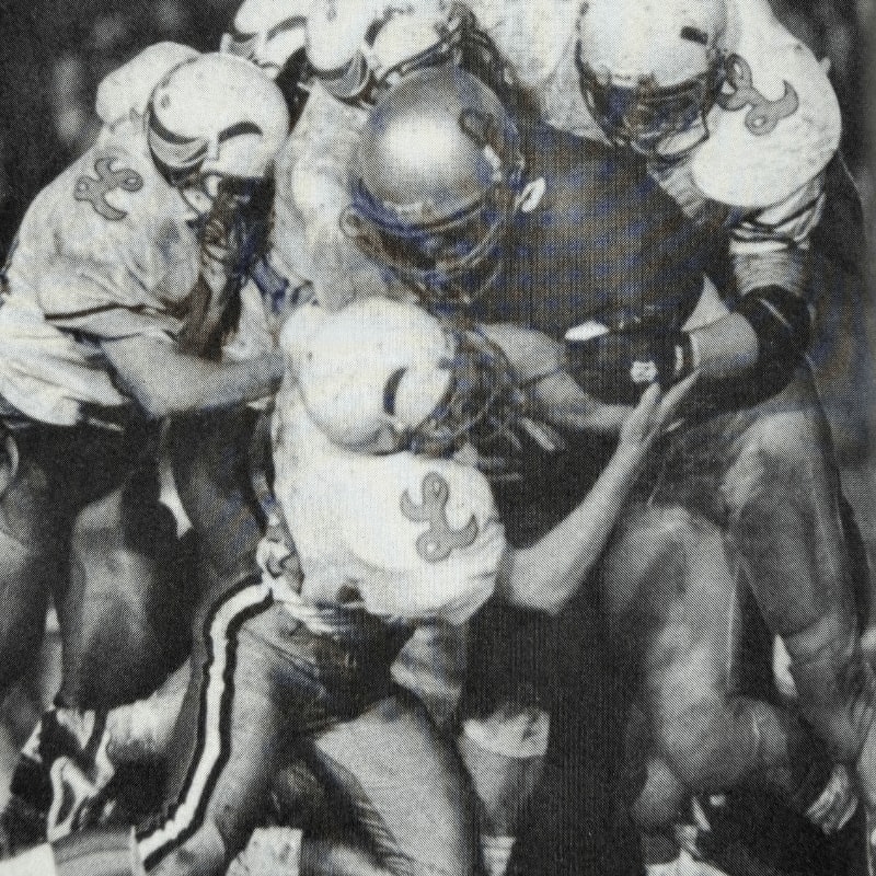 A chaotic pile of football players in uniform, some in light jerseys with a "L" logo and others in dark jerseys, are engaged in a tackle on a blurred, dark background.