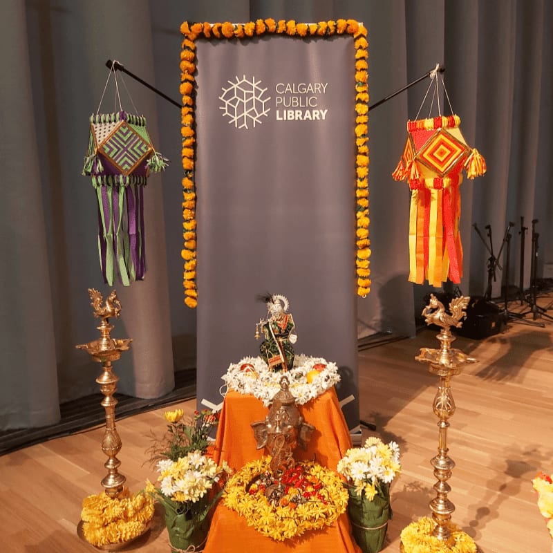 In the foreground, a colorful altar with statues and flowers is set up, while the background features a banner for the Calgary Public Library and decorative lanterns.