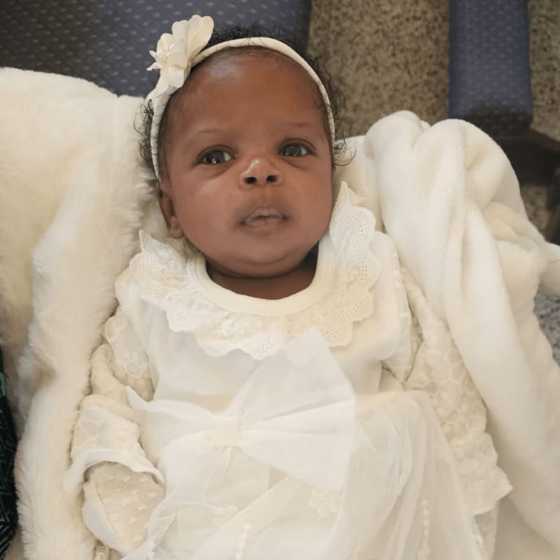 A newborn baby girl in a white christening gown and headband lies on a white fluffy blanket, with blurred blue chairs in the background.