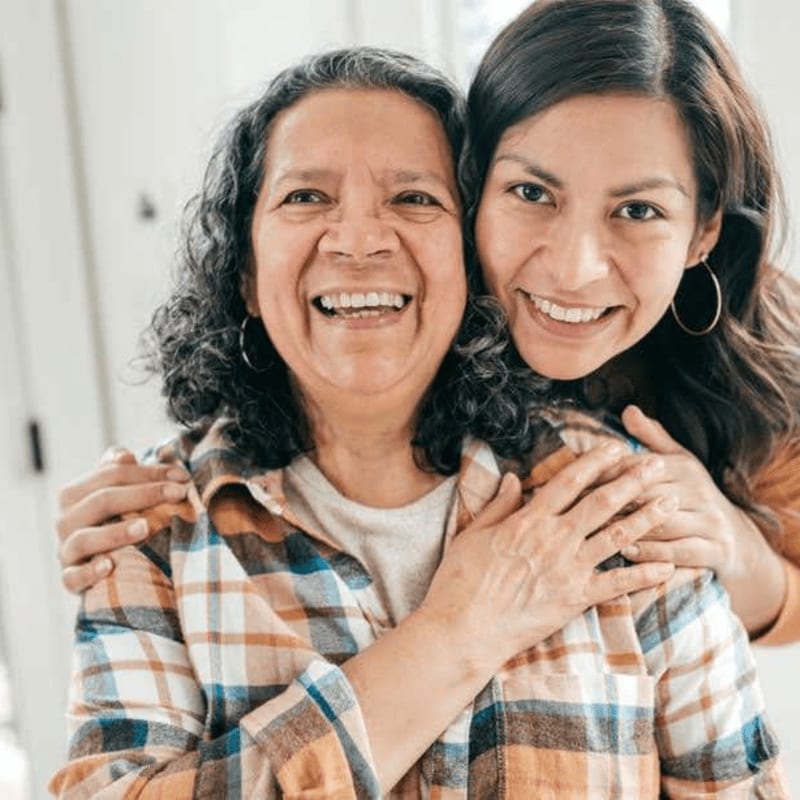 A smiling mother and daughter embrace in the foreground, with a softly blurred background suggesting an indoor setting.