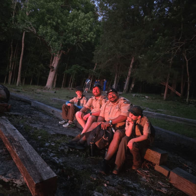 Four young men in scout uniforms sit on logs in the foreground, with a forest and a few other people visible in the dusky background.