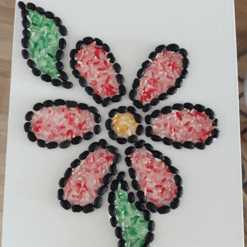 A colorful flower made of rice and beans is centered on a white canvas, with a wooden surface visible in the background.