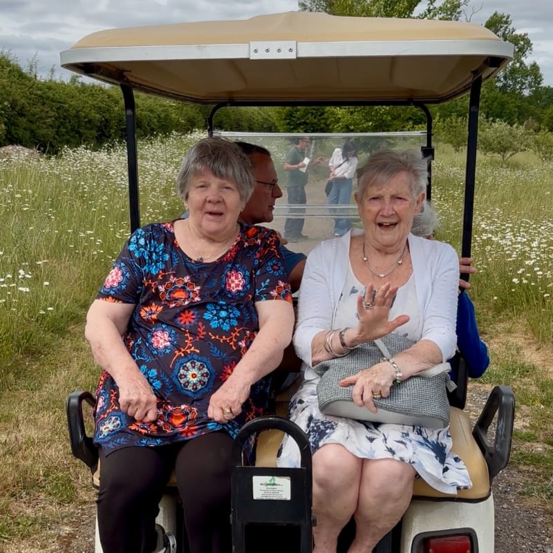 Two smiling women ride in a golf cart through a field of wildflowers, with other people visible in the distance.