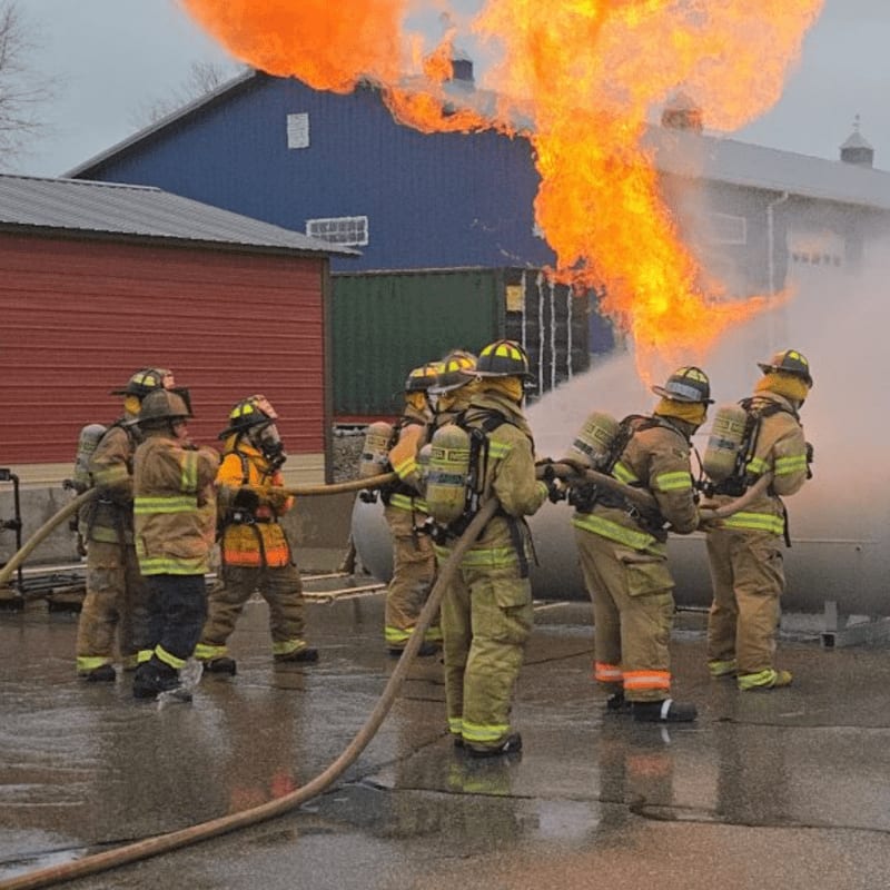 Firefighters in protective gear spray water on a large flame in front of blue and red buildings.