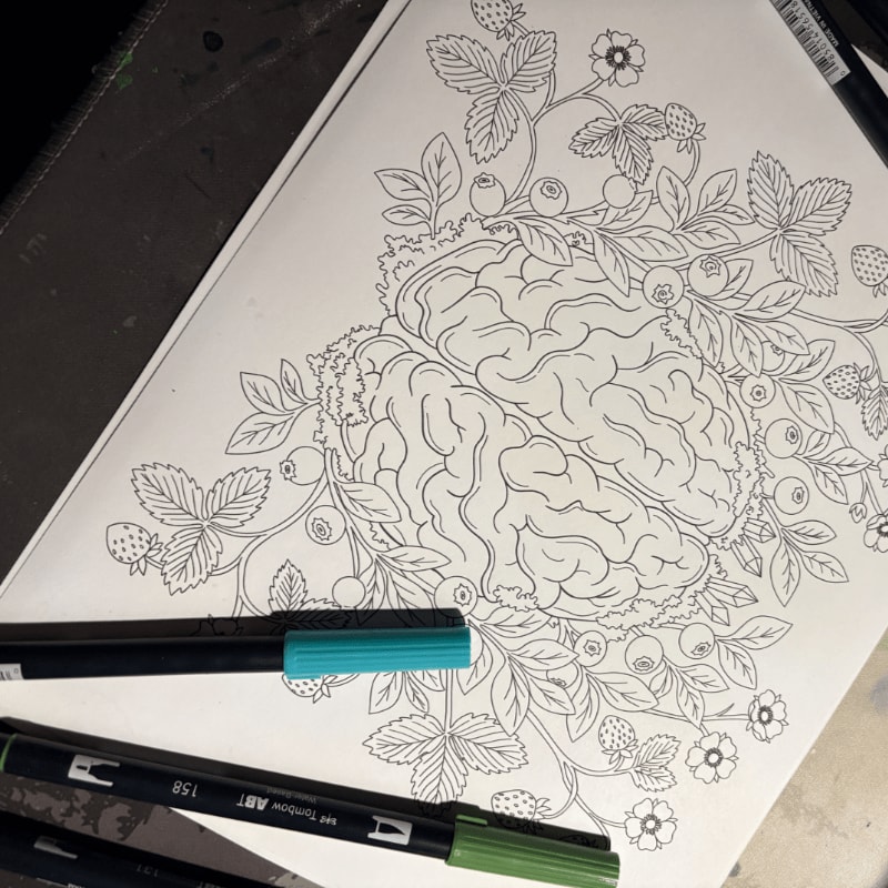 A detailed line drawing of a brain intertwined with strawberries and blueberries sits on a coloring book page, with two markers in the foreground.