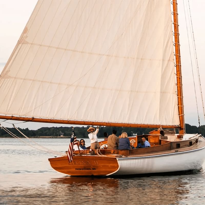 A wooden sailboat with a large white sail carries several people across calm water, with a tree-lined shore visible in the distance.