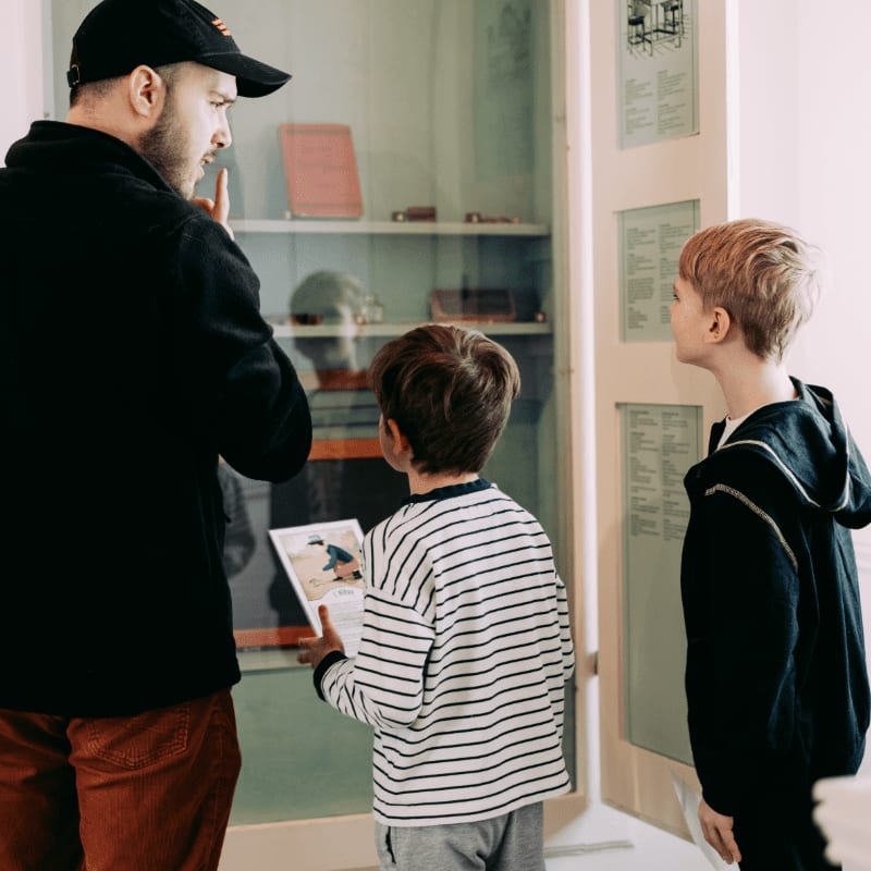 A man in a black cap and red pants points to a display case filled with artifacts, while two young boys in striped shirts and hoodies look on with interest.