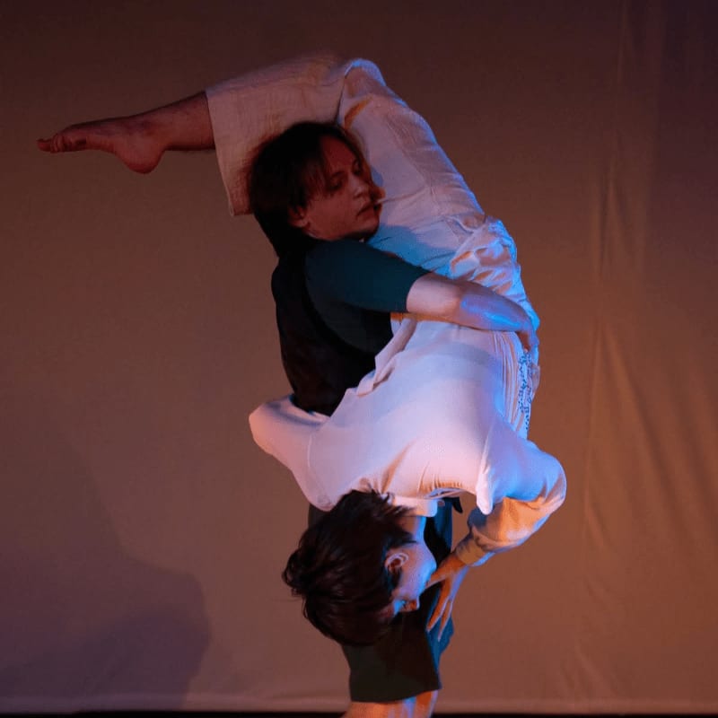 Two dancers in white clothing perform an acrobatic lift against a softly lit, plain background.