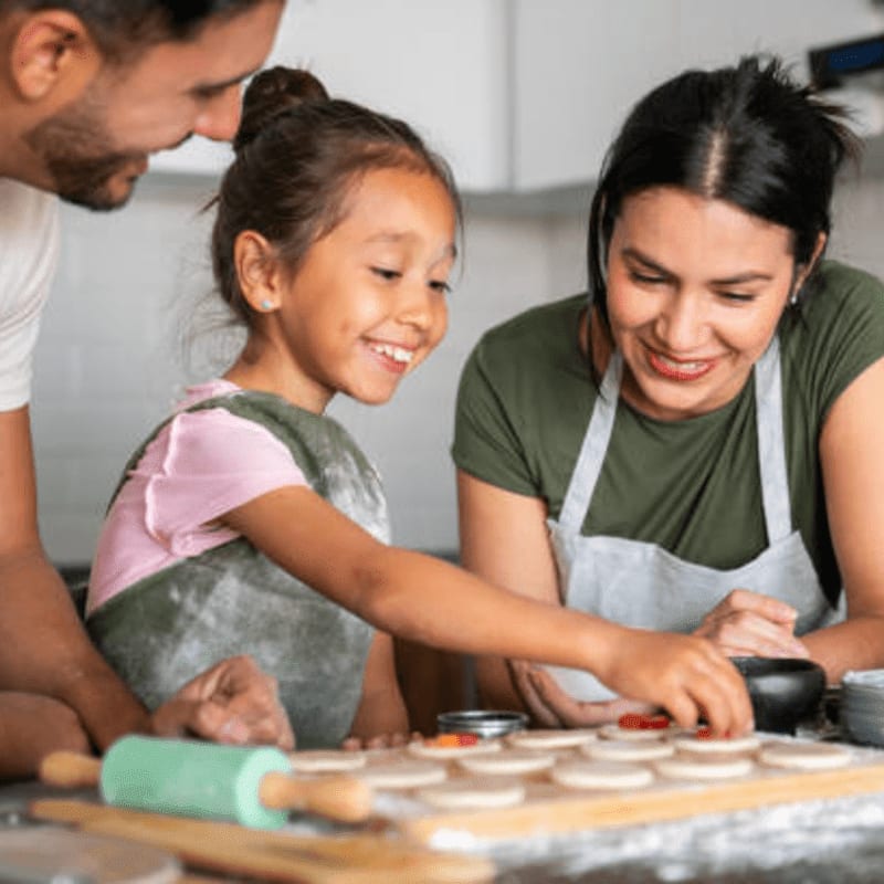 A father, mother, and daughter are happily baking cookies in a kitchen, with a rolling pin and cookie dough in the foreground and kitchen counters in the background.