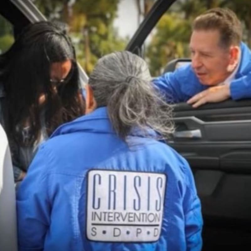 In the foreground, a person wearing a blue jacket with a "CRISIS INTERVENTION S.D.P.D." patch is seen from behind, while in the background, a man leans into a vehicle and speaks to someone.