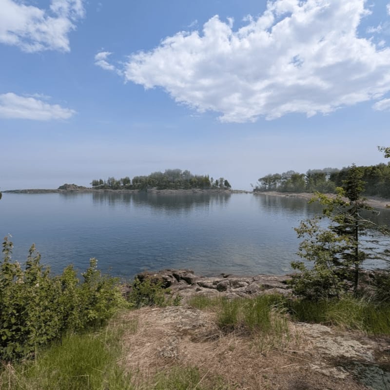 Green bushes and rocky ground fill the foreground, leading to a calm blue lake with tree-lined islands and a bright sky in the background.