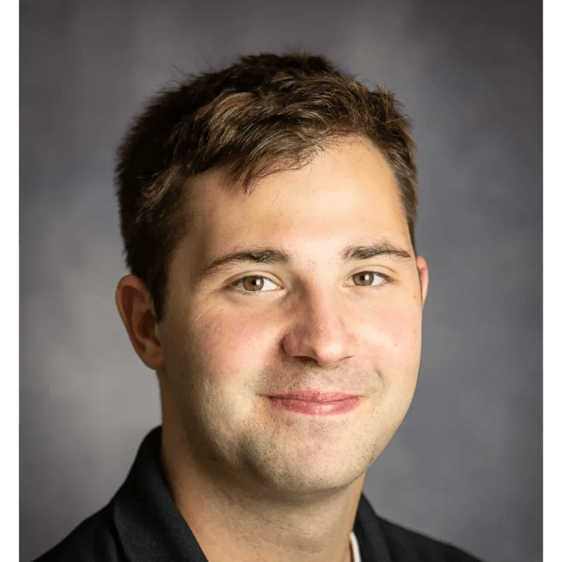 A young man with short brown hair and a slight smile is in the foreground, with a dark, textured background behind him.