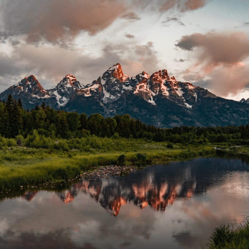 A calm river in the foreground reflects the snow-capped peaks of the Teton Mountains, which are framed by a lush green forest under a dramatic, cloudy sky.