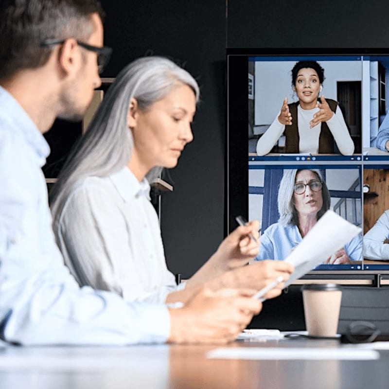 Two people in the foreground review documents while a video conference displays multiple participants on a large screen in the background.