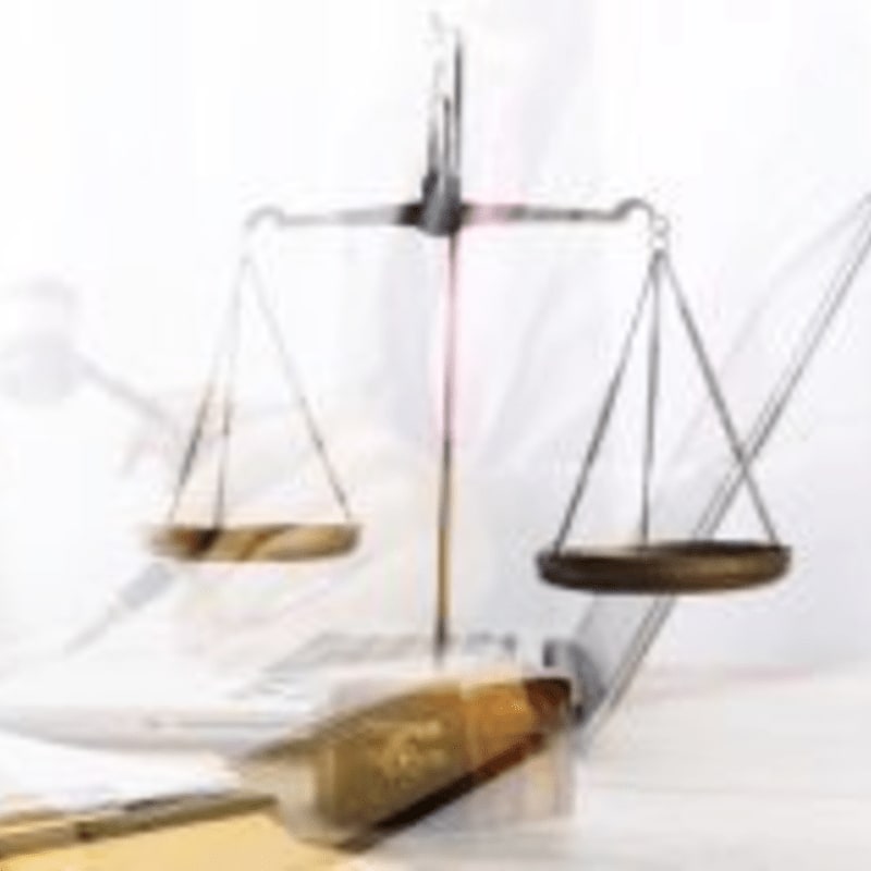 A brass balance scale rests on a stack of books in the foreground, with a blurred background suggesting a courtroom or legal setting.