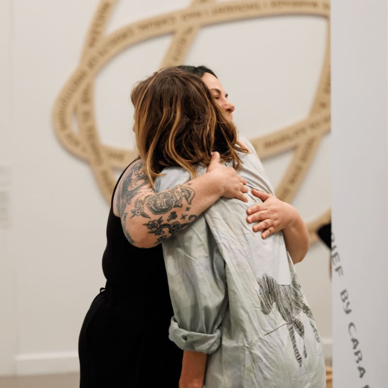 Two women embrace in the foreground, with a large, intricate artwork on the wall behind them.