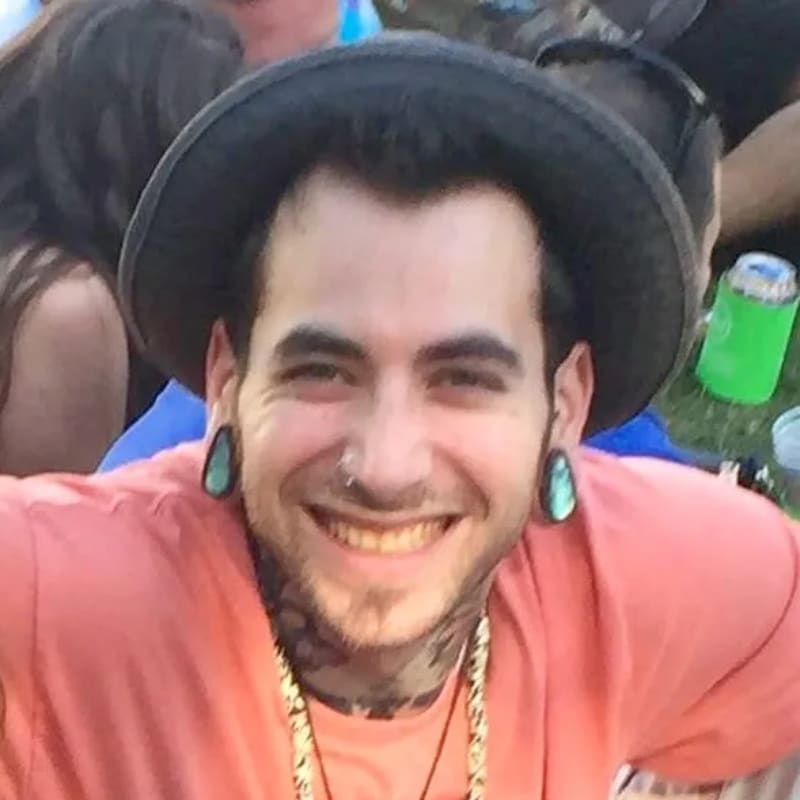 A smiling man with gauges in his ears and tattoos on his neck wears a coral shirt and a hat, with a blurred crowd in the background.