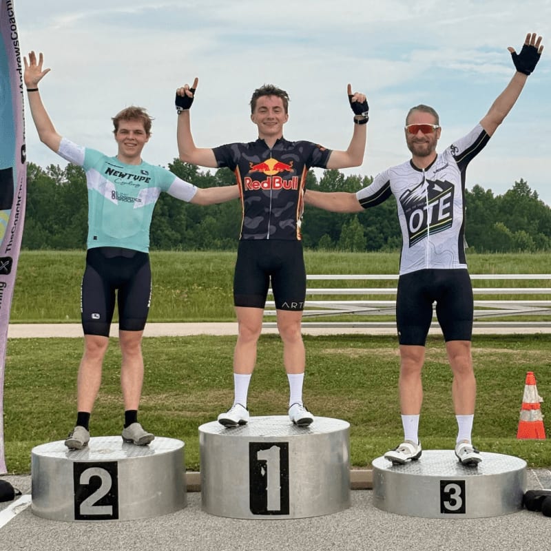 Three cyclists stand on a podium with their arms raised, celebrating their victory against a backdrop of green grass and trees under a cloudy sky.