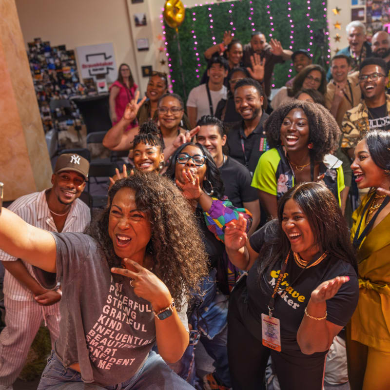 A group of smiling people, some posing for a selfie in the foreground, are gathered in a brightly lit room with a green wall and decorations in the background.