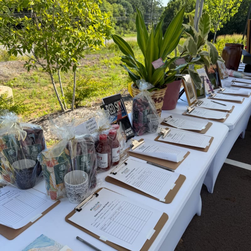 In the foreground, a white-covered table displays gift baskets, bottles, and clipboards with papers, while the background features lush green trees and a bright sky.