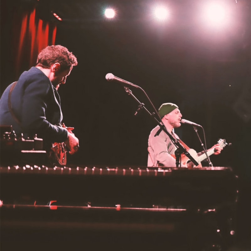 Two musicians perform on a stage with a piano in the foreground and red curtains and spotlights in the background.