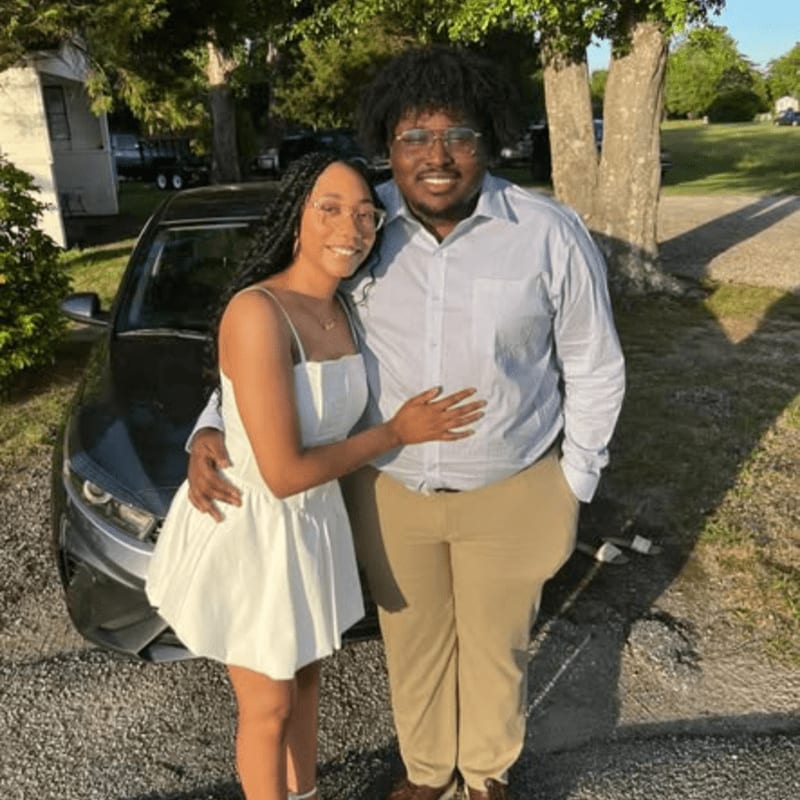 A young couple poses in front of a car with a tree and a grassy yard in the background.