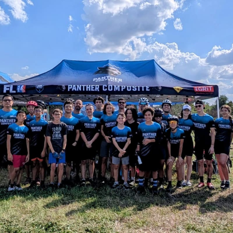 A group of cyclists in matching shirts stand in front of a tent with a blue sky and clouds in the background.