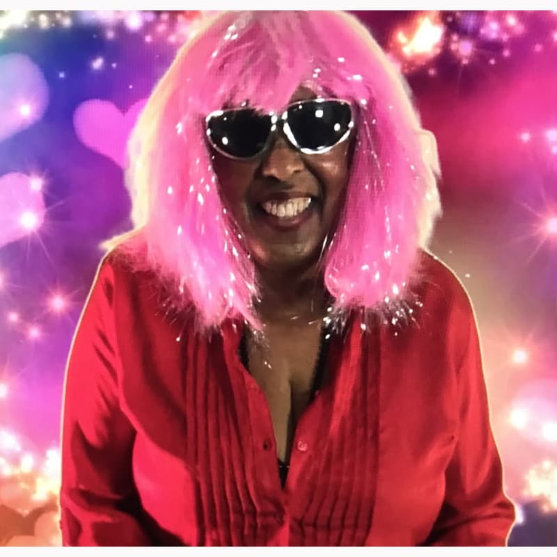 A smiling woman in a bright red shirt and a pink wig with sparkly accents wears sunglasses against a background of glowing hearts and lights.