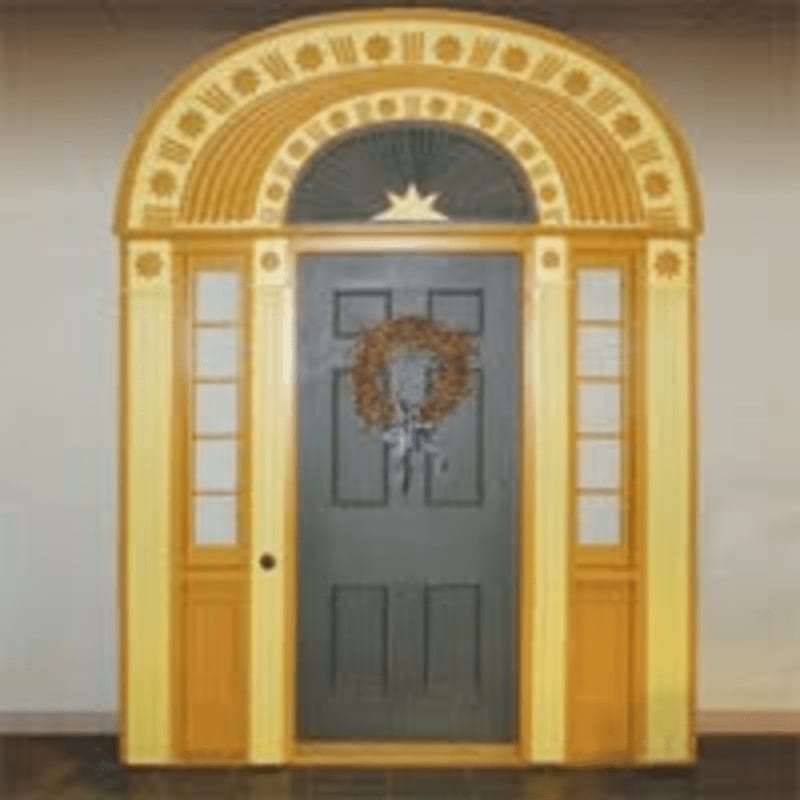 A dark gray door with a wreath hangs in the center of a yellow arched entryway with sidelights, set against a plain background.
