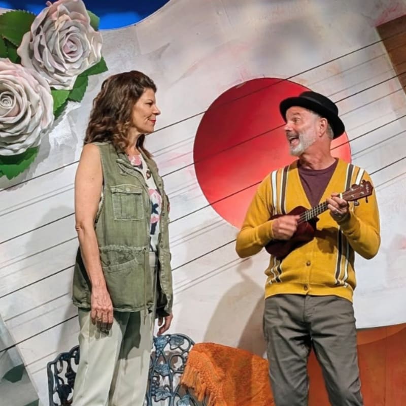 A woman in a green vest and a man in a yellow cardigan play a ukulele on a stage with large white roses and a red circle in the background.