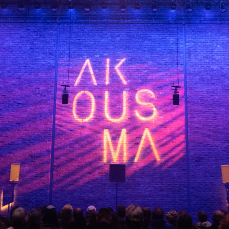 A crowd of people watches a stage with a brick wall illuminated by blue and pink lights, displaying the word "AKOUSMA" in neon letters.