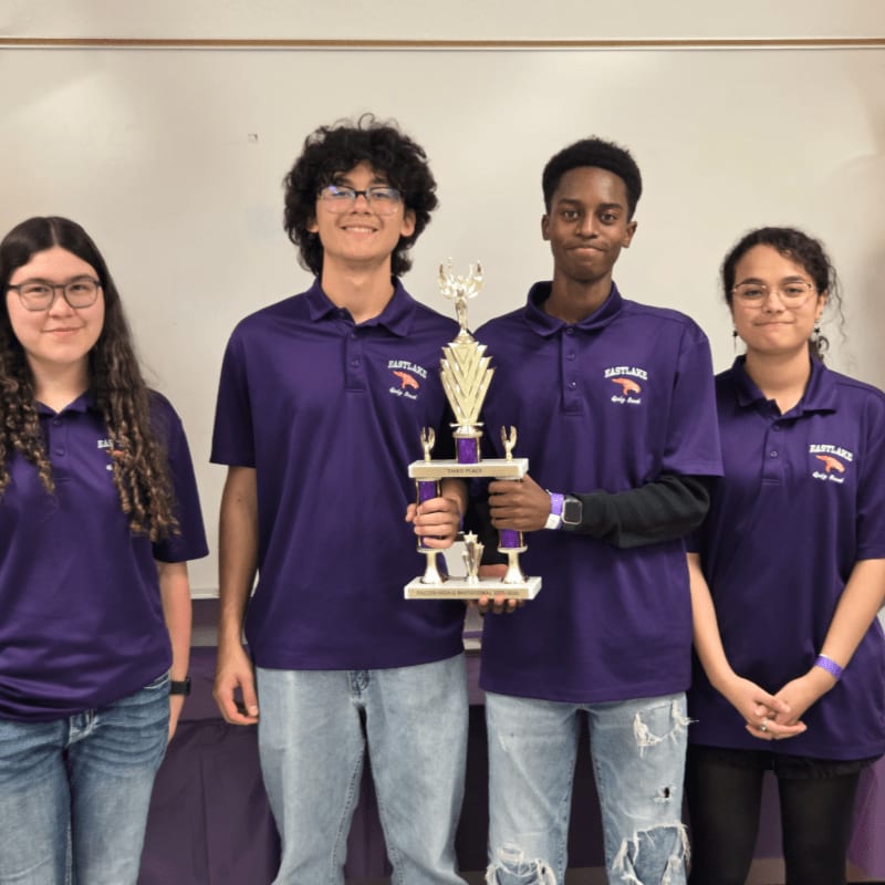 Four smiling teenagers in purple shirts stand in front of a white wall, holding a trophy.