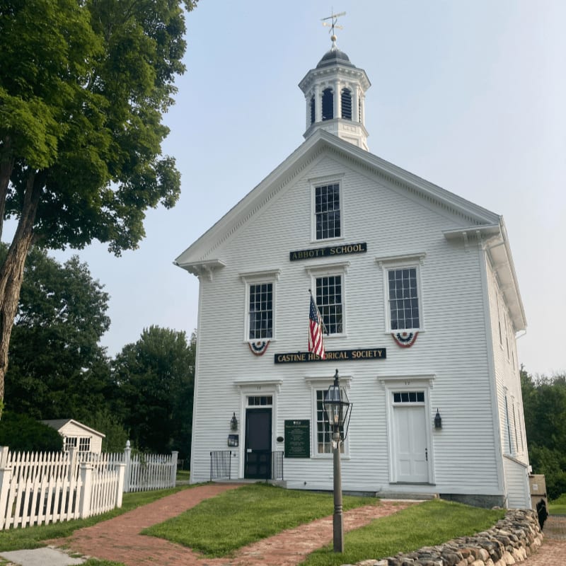A white historic schoolhouse with a cupola stands on a grassy lawn with a white picket fence in the foreground, against a clear sky.